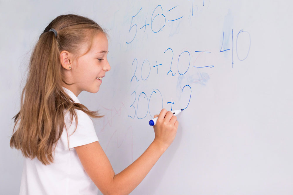 Mädchen schreibt mathematische Aufgaben mit blauem Stift an eine weiße Tafel