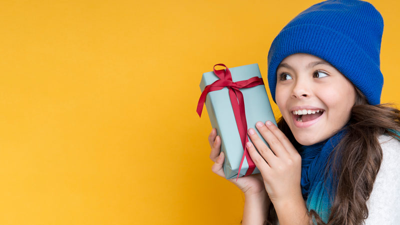 Mädchen mit Mütze hält Geschenk vor gelbem Hintergrund